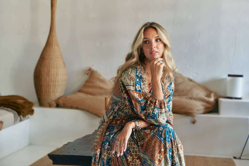 Teresa Maxi Dress in Izabelle Print featuring bohemian brown and blue patterns, worn by a woman seated in a minimal, earthy-toned room.